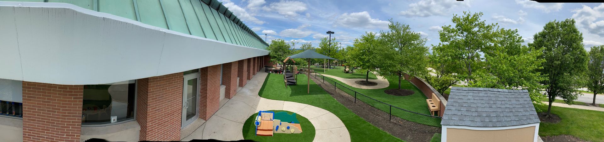 A panoramic outdoor view of a brick building with a green roof, a grassy courtyard, trees, and a small shed.