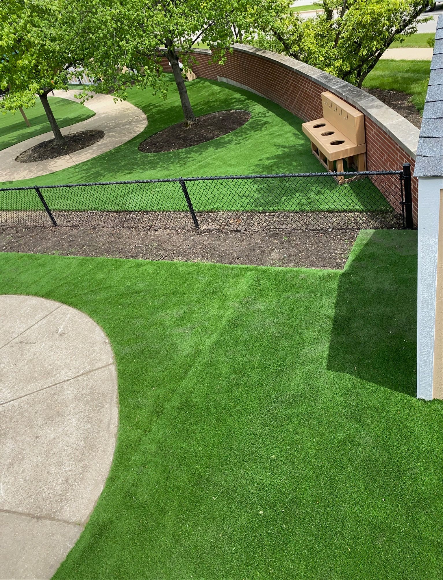 A view of a grassy yard with a curved brick wall, a small wooden bench, trees, and a concrete path.