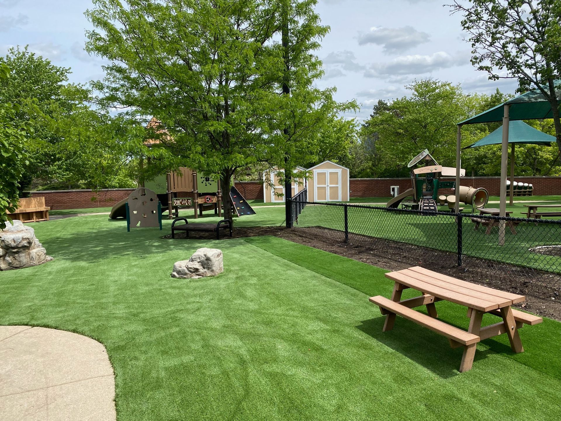 A playground with green artificial turf, a wooden picnic table, climbing structures, a small shed, and trees.