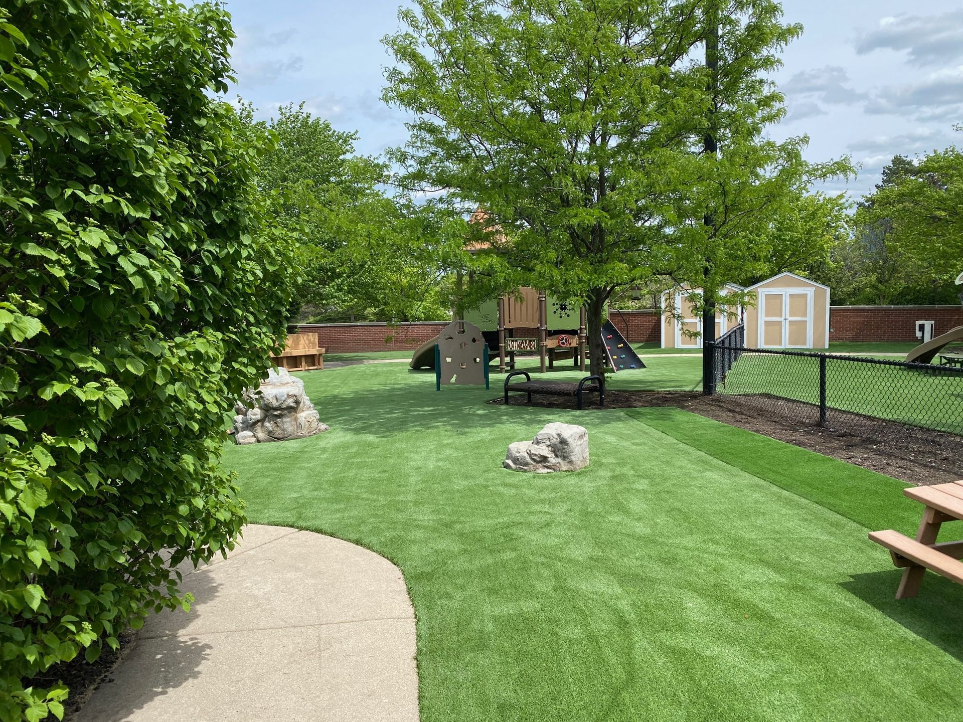 A playground with artificial grass, climbing rocks, play equipment, and a wooden picnic table under a large tree.