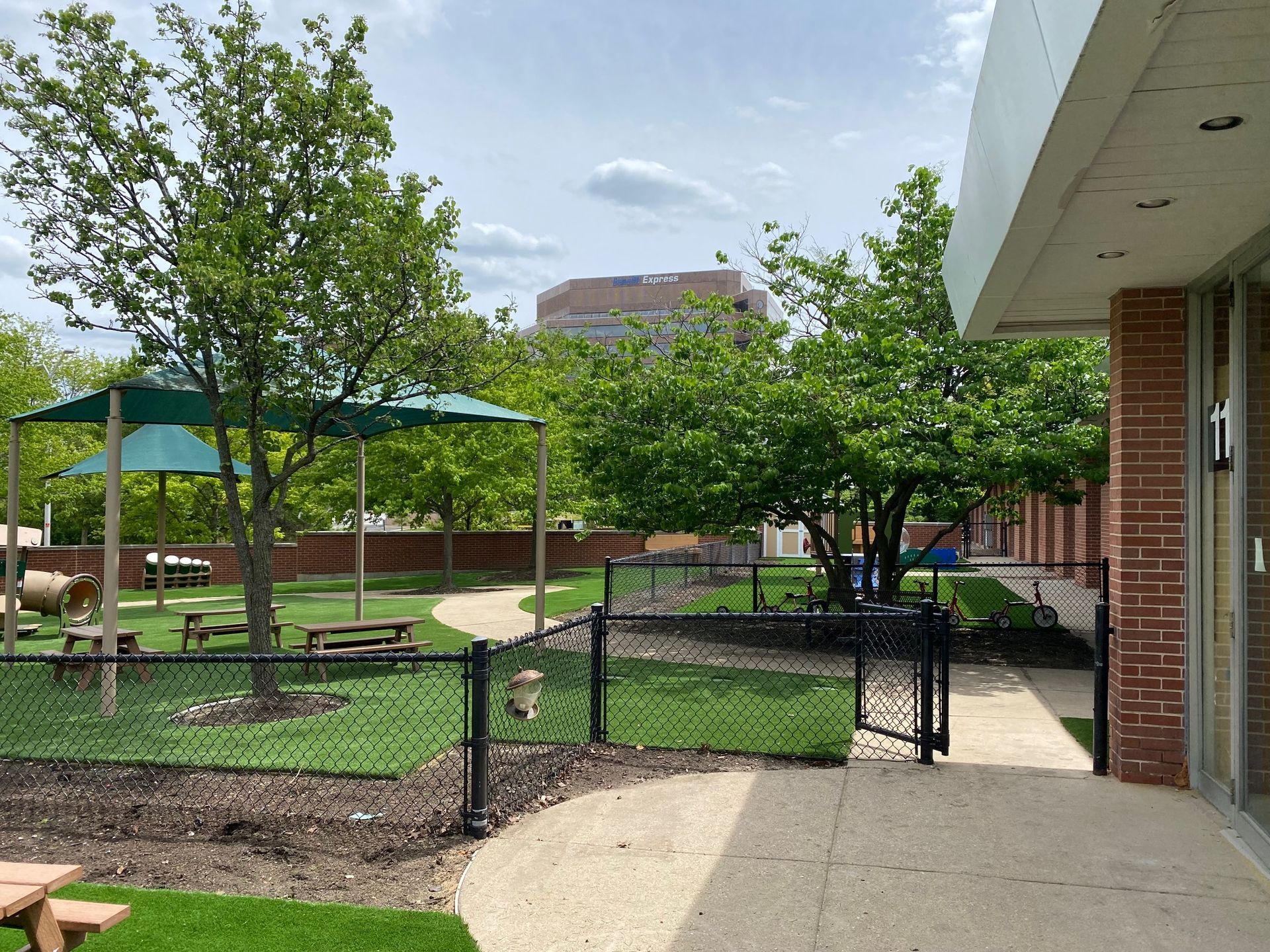 A playground area with a green shade canopy, picnic benches, and trees, enclosed by a black chain-link fence.