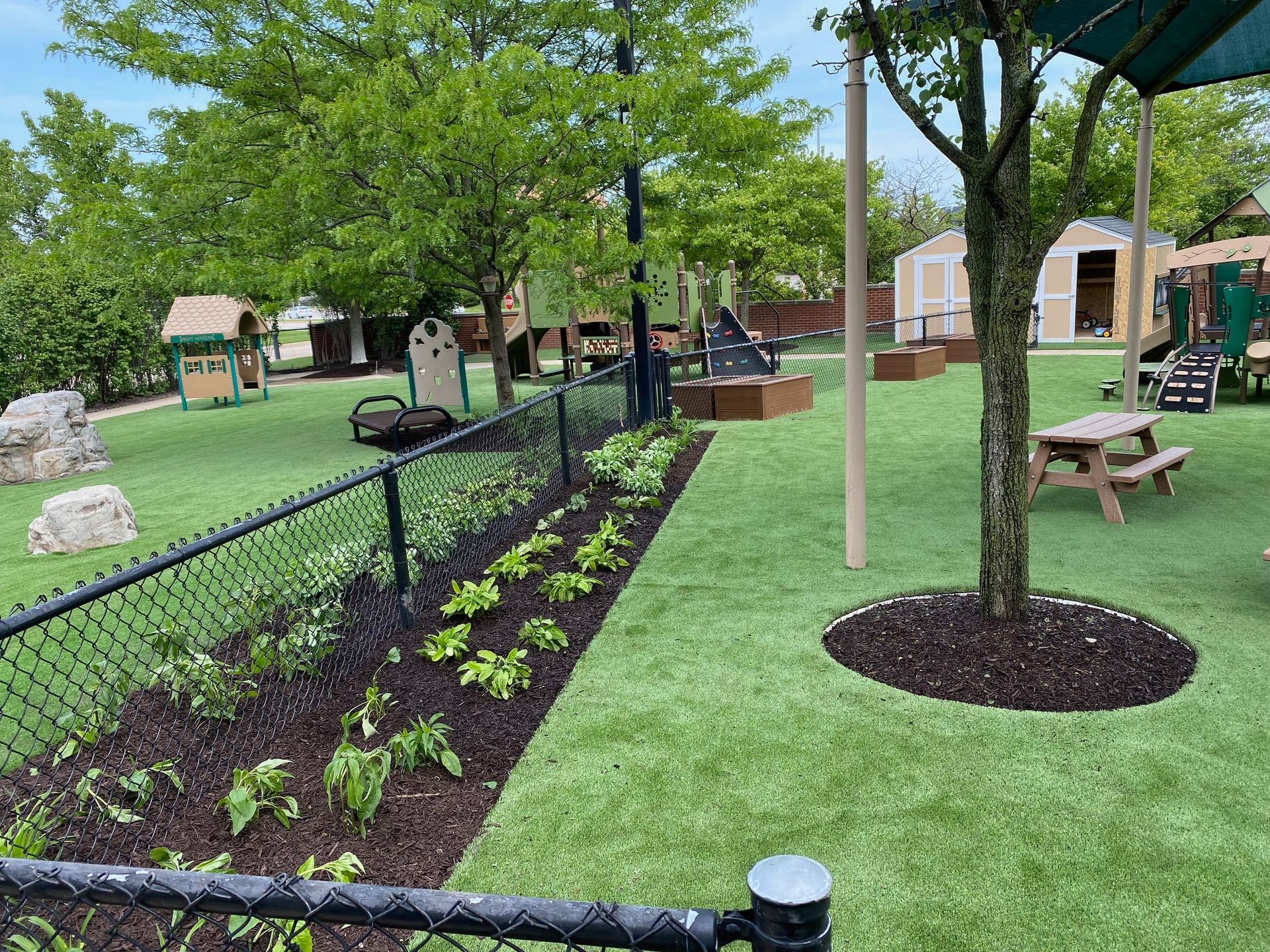 A green playground featuring a chain-link fence, garden beds with mulch, a picnic table, and play equipment on a sunny day.