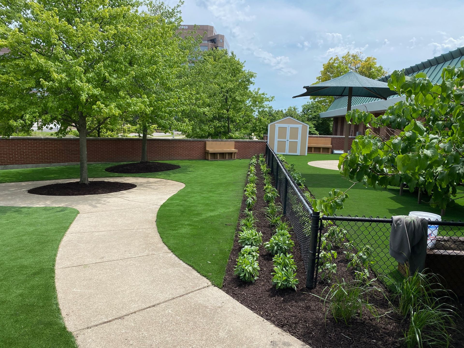 A landscaped park with a winding gravel path, green grass, young trees, a garden bed, and a shed in the background.