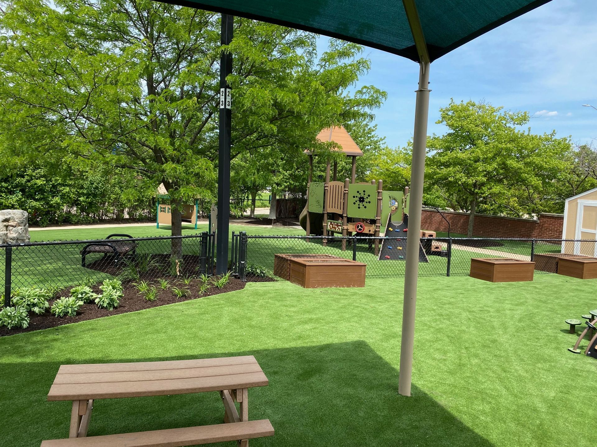 A sunny playground with a wooden picnic table, green lawn, garden beds, and a play structure under a shade canopy.