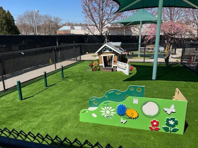 Playground with green turf, playhouse, and educational wall against a black fence.