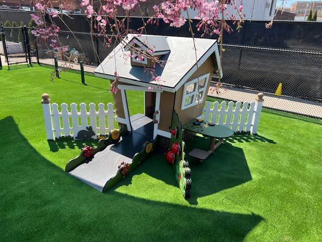 Playhouse with picket fence on artificial grass under a blooming tree.