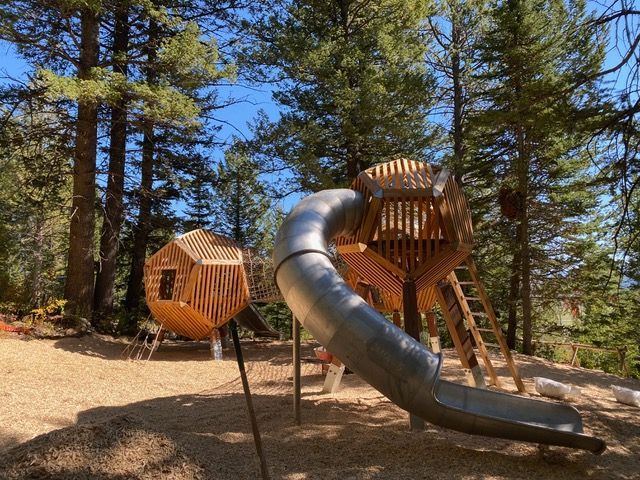 Playground with wooden structures, metal slide, and wood chip ground in a forest setting.