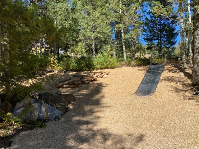 Playground with wood-chip ground cover, log structures, and a wooden ramp in a forested area on a sunny day.