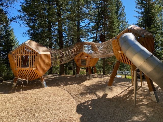 Wooden playground with treehouse structures, net bridges, and a metal slide, set in a woodchip-covered area.