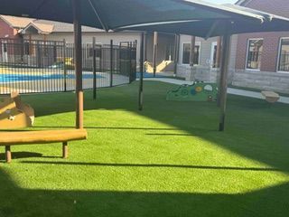 Playground with artificial turf, shade structure, and pool in background.
