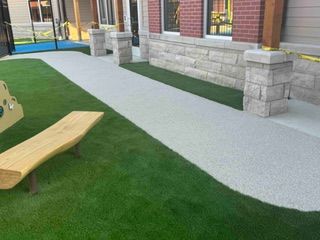 Pathway of light-colored aggregate concrete with a green lawn and a wooden bench. Brick building in the background.