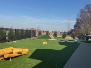 A grassy park with play equipment: logs, a hill, and benches. Fenced area under a blue sky.