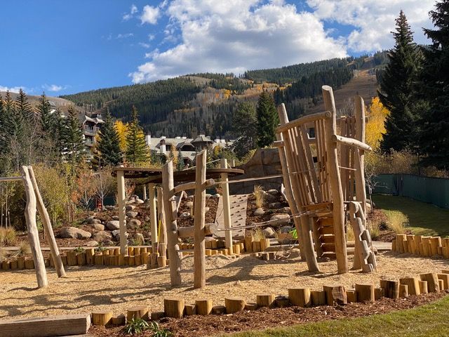 Wooden playground with climbing structures, sand, and mountain backdrop.