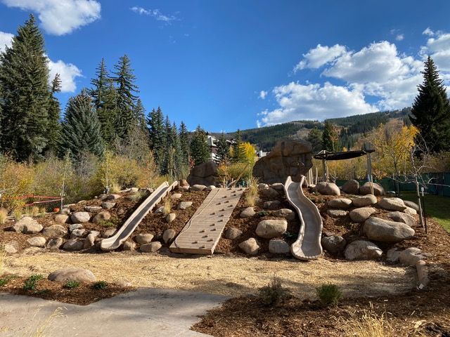 Playground with two slides, climbing wall, and large rocks, under a blue sky.