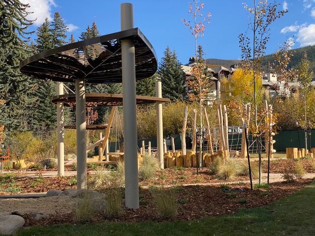Playground with wood structures, trees, and foliage under a blue sky, mountain background.
