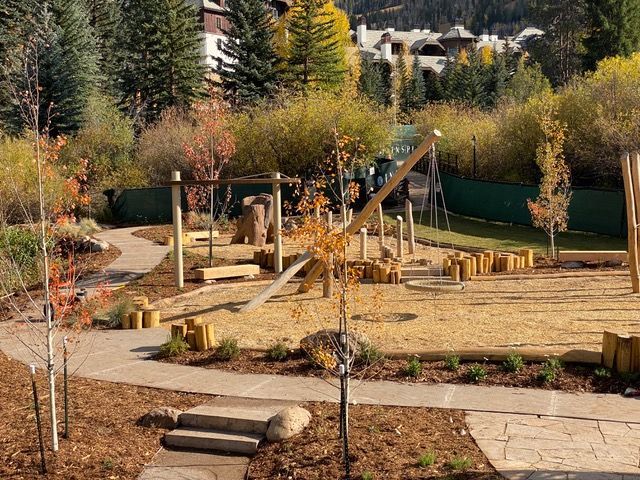 Playground with wood structures, swings, and climbing logs surrounded by trees and a walking path.
