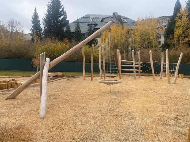 Wooden playground with climbing structures and swings, surrounded by wood chips, trees, and buildings.