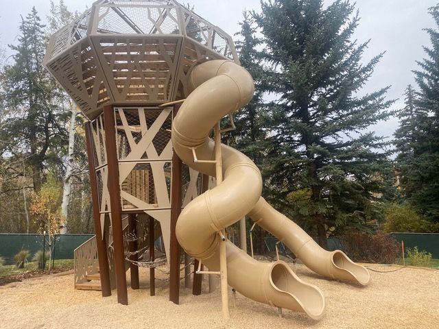 Playground with beige tower, twisting slide, and tan ground. Trees in background.