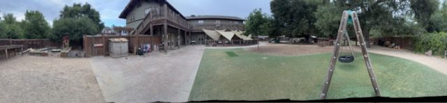 Playground with a swing set and a building with tents in the background. Green grass and overcast sky.