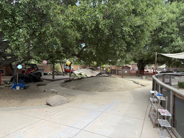 Playground with trees, dirt area, and play structures. Concrete path with blue lines.