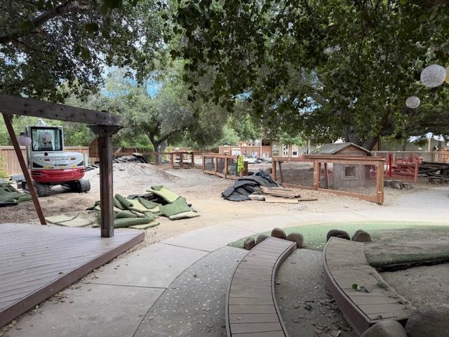 Playground under construction; small excavator, dirt, wood structures, green turf, trees, and walkway.