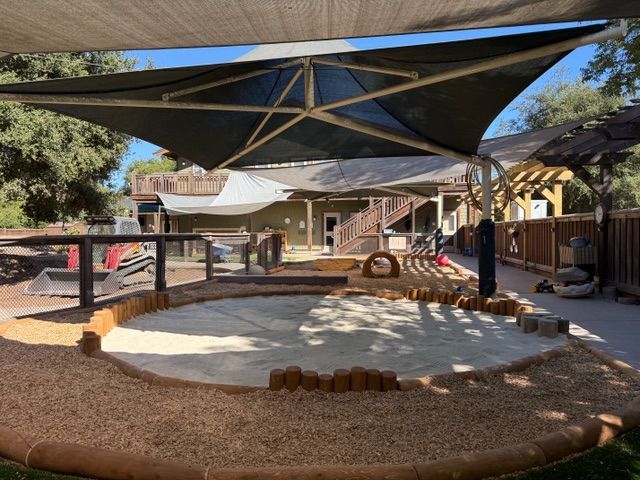 Outdoor playground with shade structure over a sandbox. Brown edging, wood chips. Building in background.