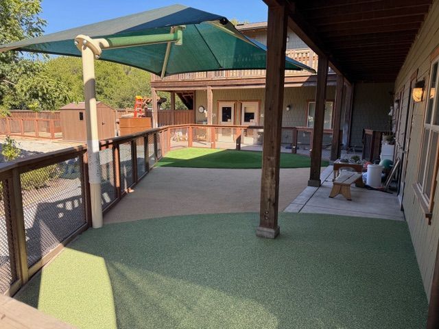 Outdoor play area with green turf, shade structure, and wooden railings.