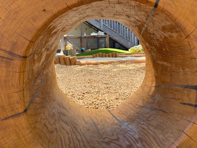 View through a wooden tunnel towards a playground with gravel, grass, and buildings.
