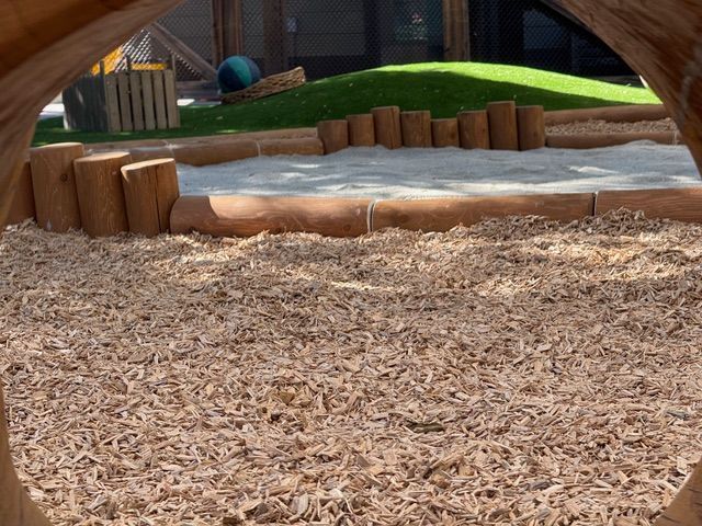 View through a wooden structure of a sandbox bordered by wooden logs, with a grassy hill in the background.