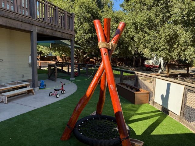 A-frame play structure with swing, on a green artificial lawn, next to a building and trees.