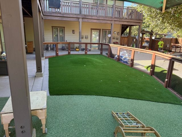 Enclosed green play area with artificial turf, brown fence, and a building with a balcony in the background.
