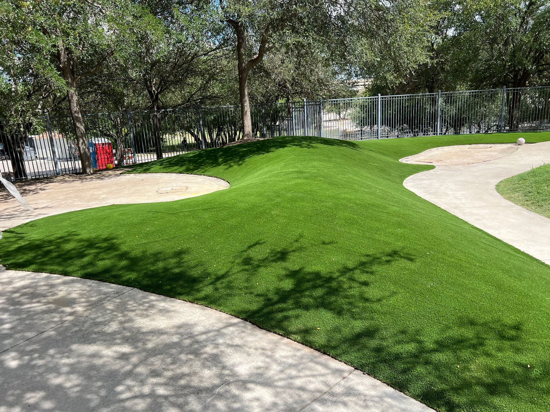 Green grassy hill in a playground with winding pathways and sand areas, trees in the background.