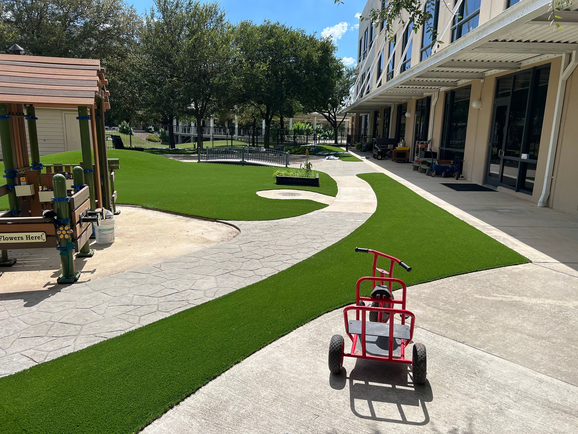 Playground with a red wagon on paved pathway, surrounded by green turf and a building.