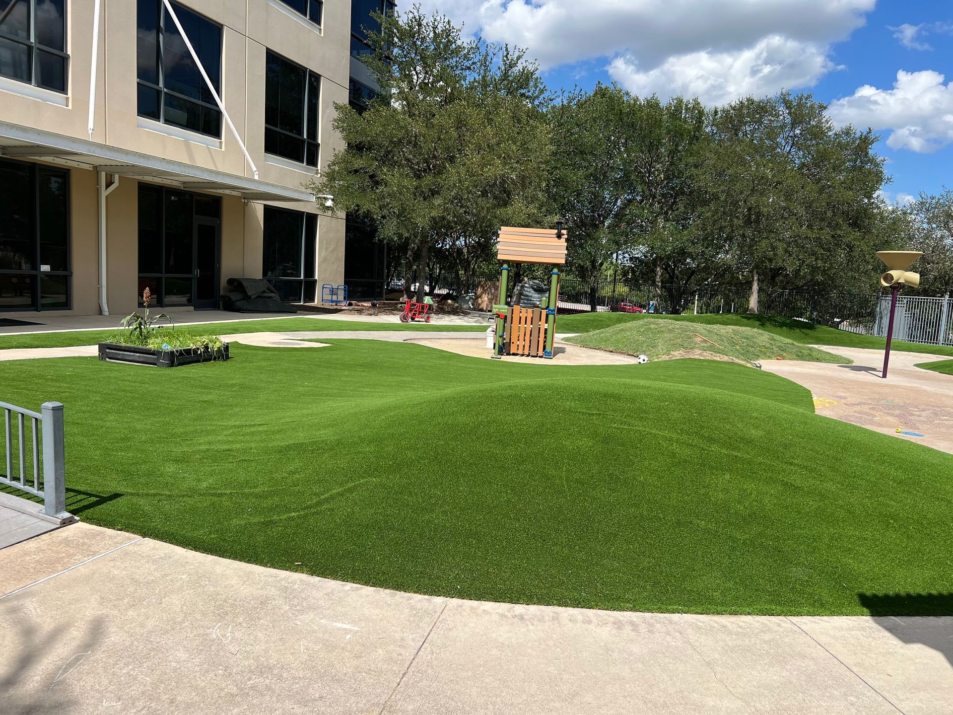 Outdoor play area with artificial turf, hills, and a wooden structure near a building.