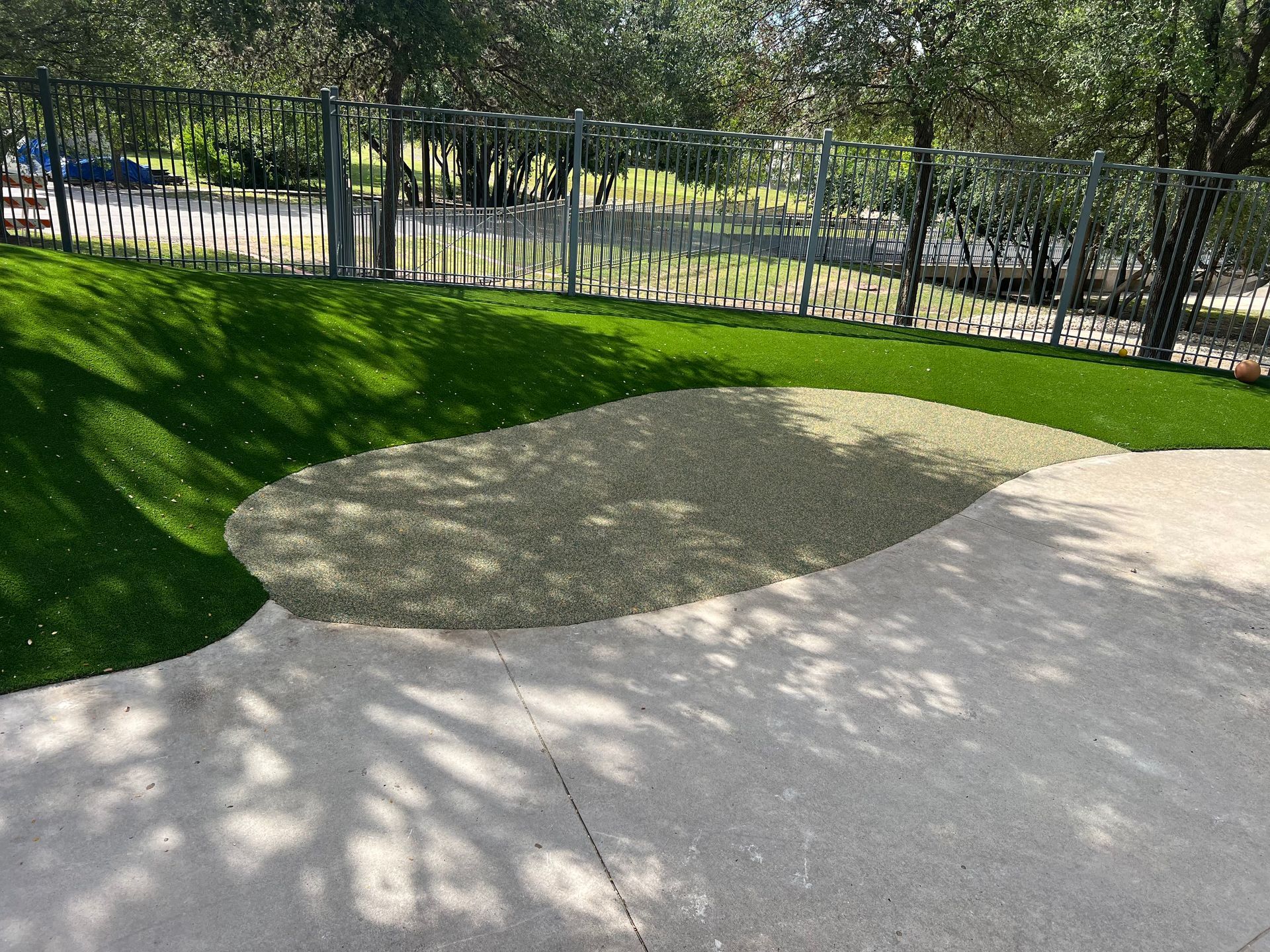 Green turf, gravel, and concrete ground area near a fence and trees.