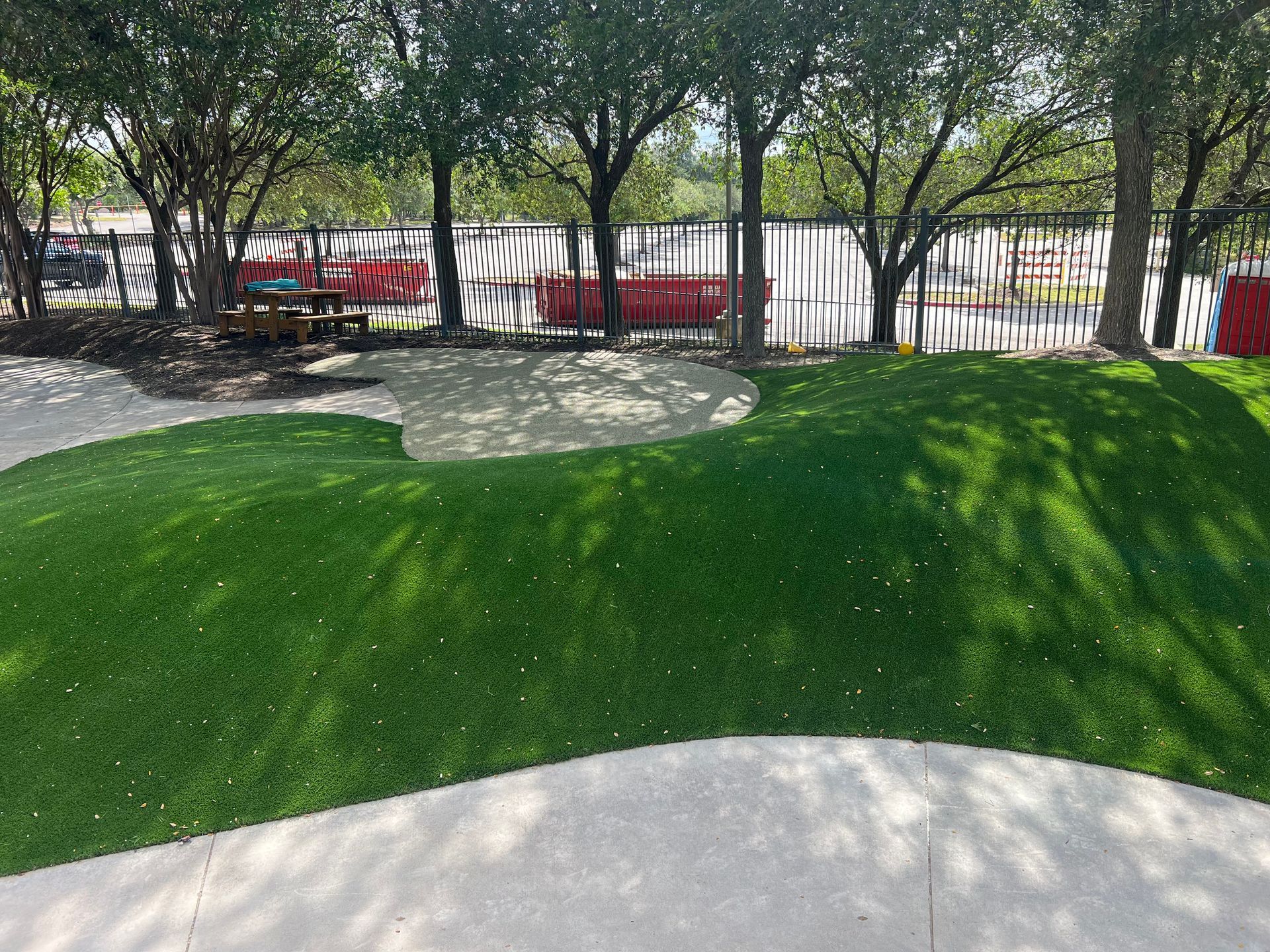Green turf playground with sand area, trees, and fence in background.