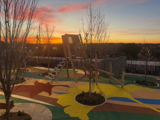 A playground with a wooden climbing structure and colorful painted ground, set against a vibrant orange sunset.