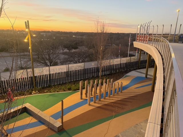 Playground at sunset with colorful ground surfacing, wooden posts, and a curved elevated walkway with metal railings.