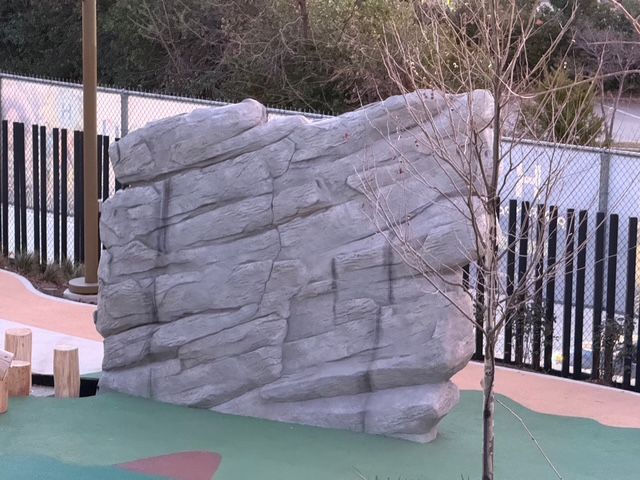 A gray, artificial rock-climbing wall stands on a green outdoor playground surface near a bare tree and metal fence.