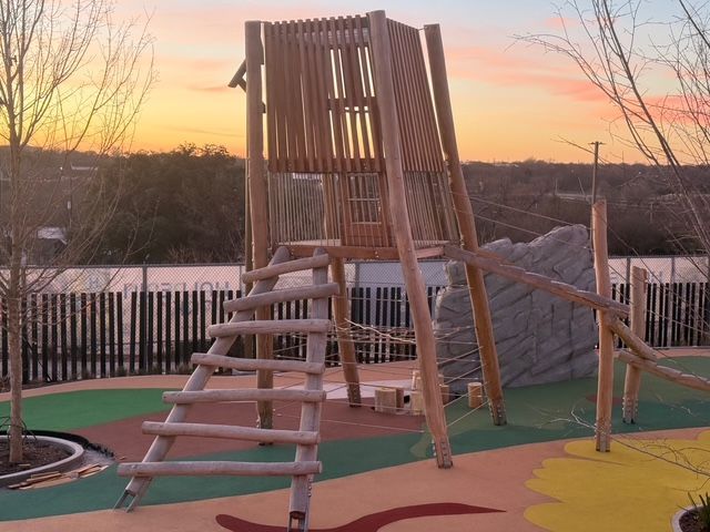 Wooden playground structure with a ladder and climbing wall on a colorful padded surface at sunset.