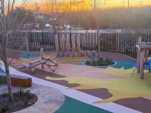 A colorful, sun-themed playground at sunset with wooden log structures, circular planters, and a wooden slat fence.