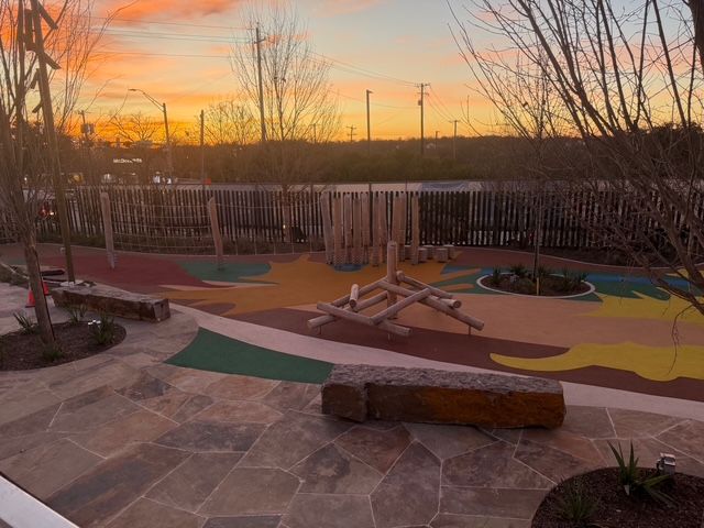 A modern playground with colorful, organic-shaped flooring, wooden structures, and stone benches at sunset.