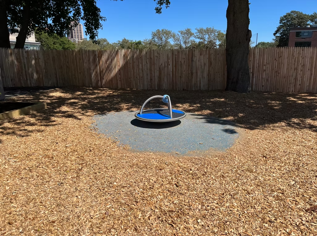 A playground with a blue spinning carousel on a gravel bed, surrounded by wood chips and a wooden fence.