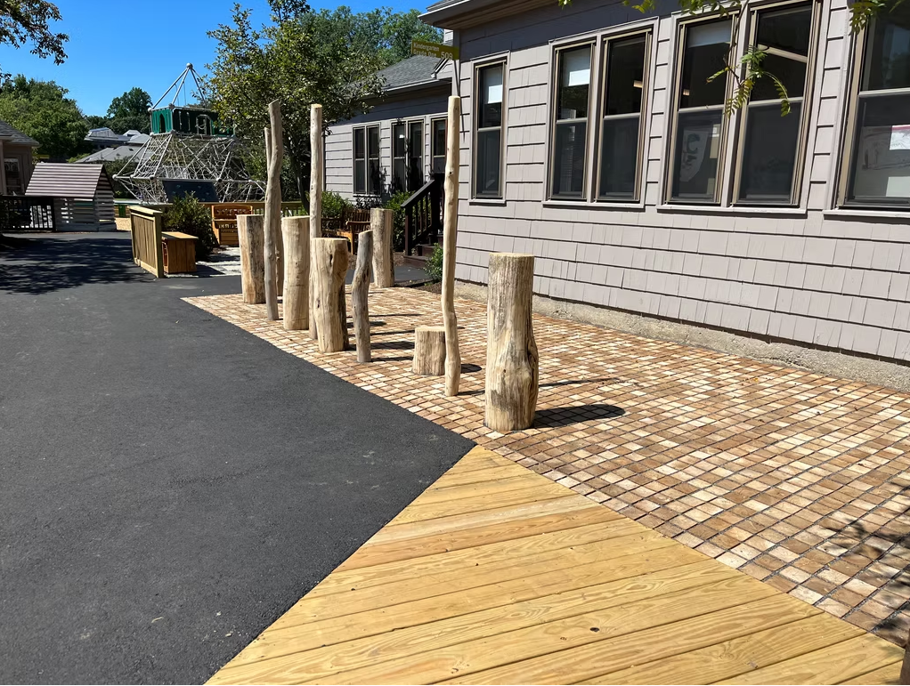 Wooden posts line a pebble path next to a building with windows, with a wooden ramp in the foreground.