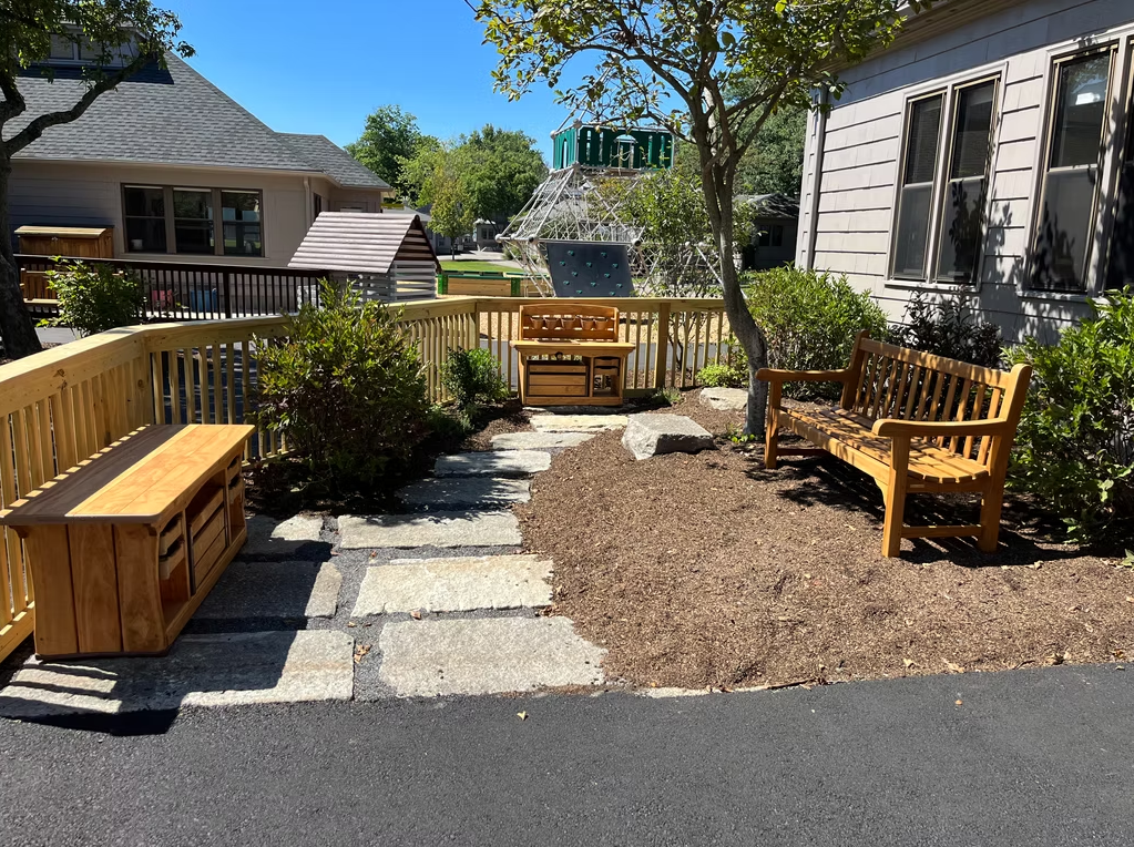 Outdoor seating area with wooden benches, stone path, mulch, and surrounding buildings.