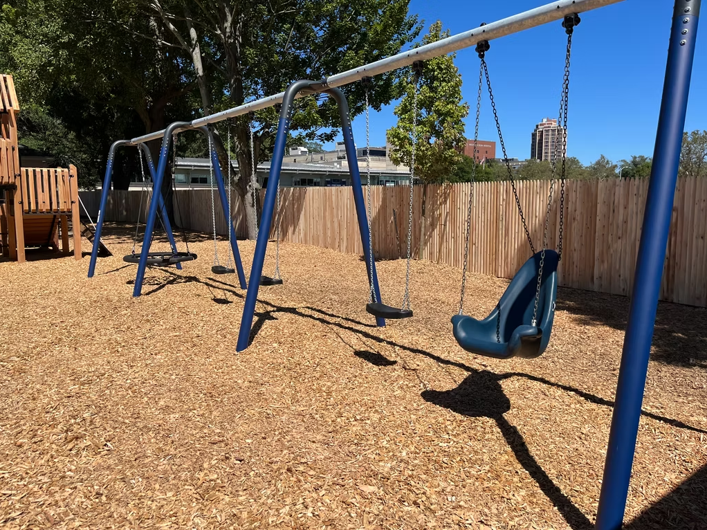 Swing set on wood chip ground, blue metal frame, sunny day.
