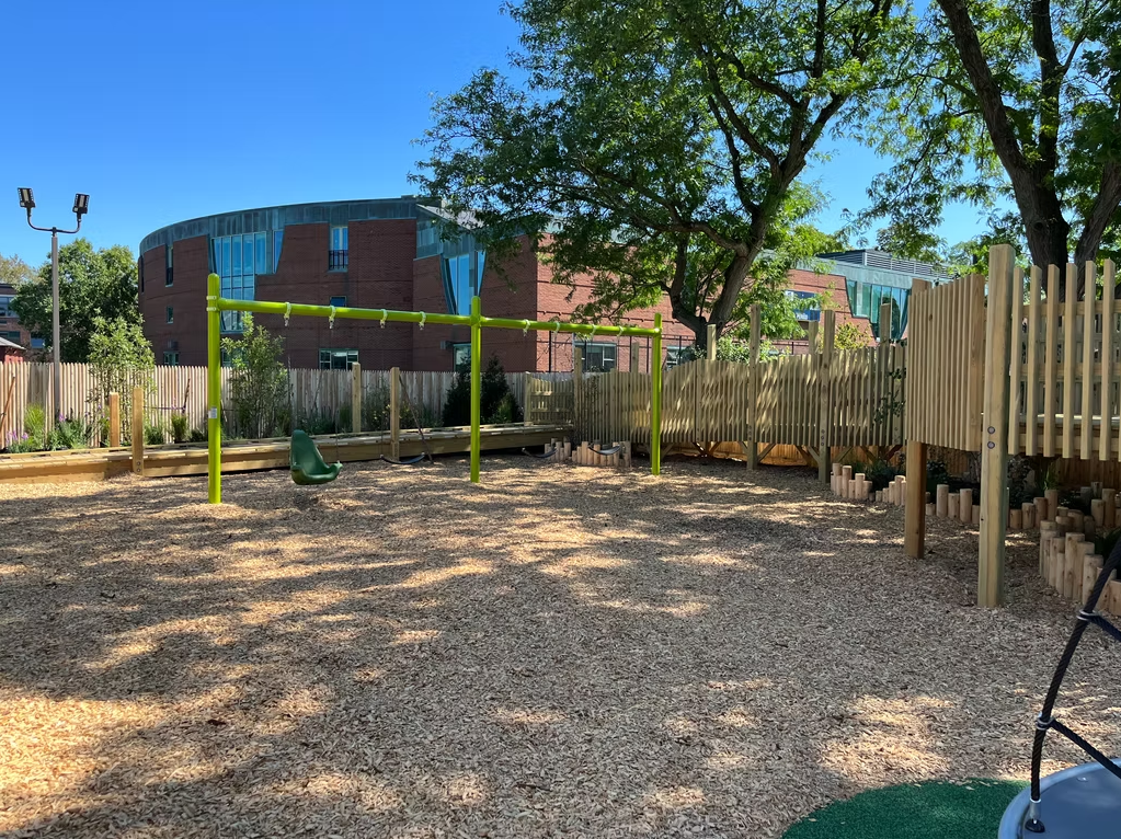 Playground with wood chip ground cover, wooden fence, and green play equipment.