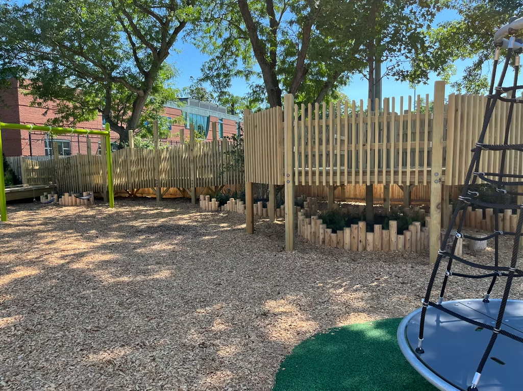 Playground with wood chip ground cover, wooden fence, and climbing structure.