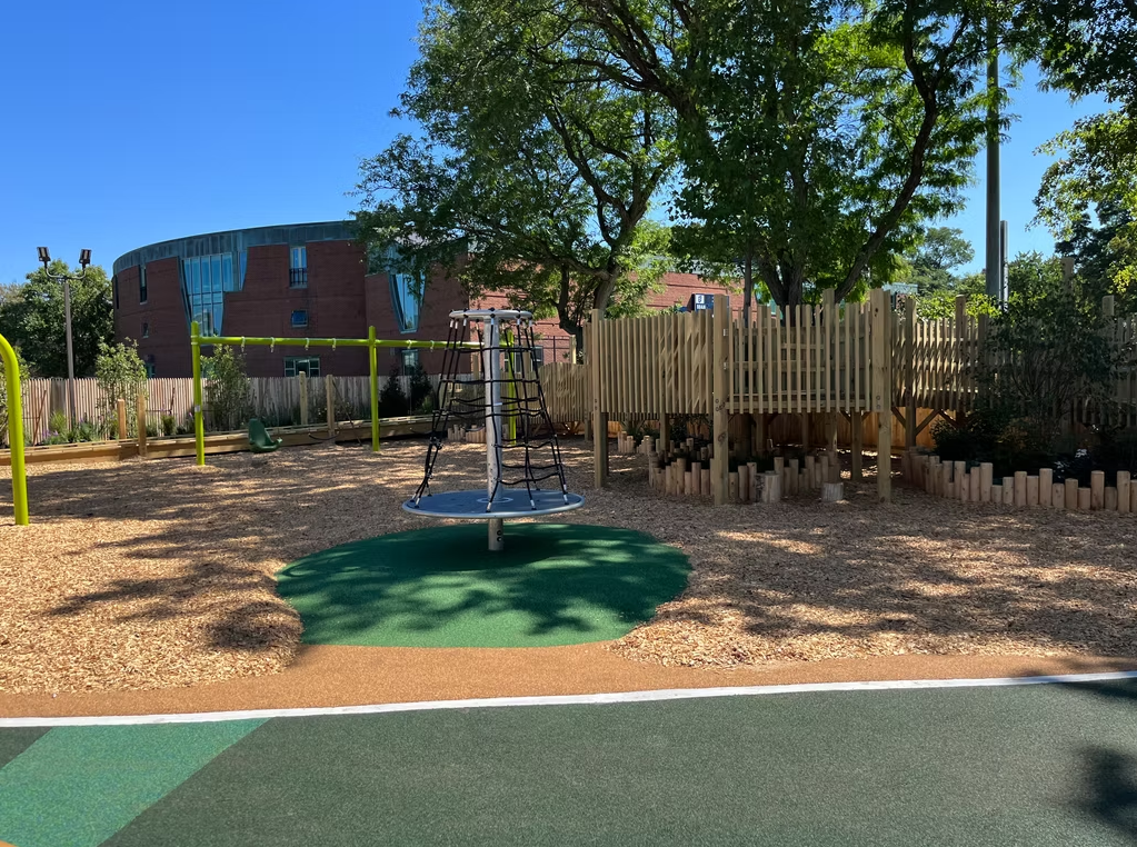 Playground with a spinning structure, wood fence, and building in the background. Brown mulch and green ground covering.