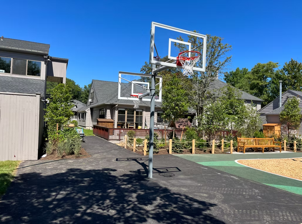 Basketball hoops on a black court in a residential area. Trees and buildings in the background on a sunny day.